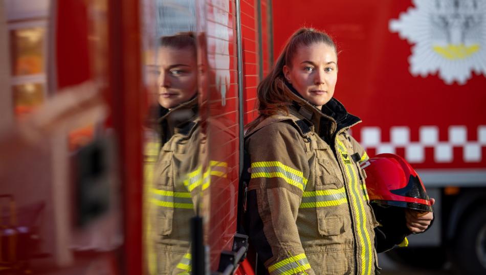 Kvinnelig brannkonstabel i utrykningsbekledning foran rød brannbil