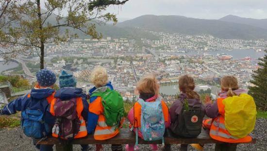 Seks barn sitter på benk med sekk på ryggen med ryggen mot kamera og ser ut over Bergen