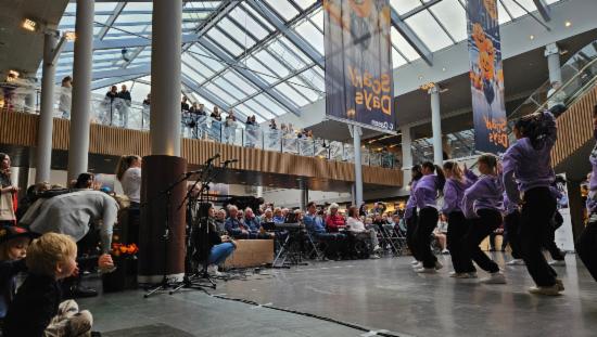 En dansegruppe framfører en dans for mange publikummere, i et stort og lyst lokale med glasstak og rulletrapp.